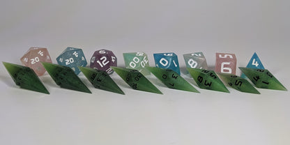 A size comparison of swirling shades of green with black numbering long pointed diamond handmade dice with standard shapes placed behind for scale. The photo emphasizes the length of the diamond dice being much greater than a standard set as well as the diamond set being much more slender. This artisan dice set features 2 d20s and rests on a seamless white background to highlight the elongated profile of the resin dice.