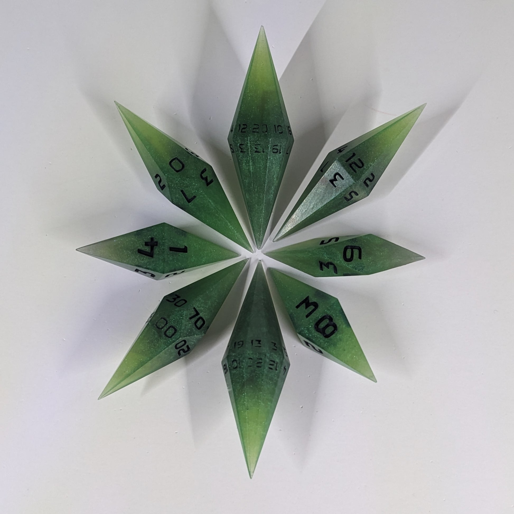A complete set of resin dice featuring 2 d20s for advantage rolls. These swirling shades of green with black numbering slender handmade diamond dice are clustered in a tight circle on a white background, highlighting the quality of the artisan dice craftsmanship and the unique pointed shape.