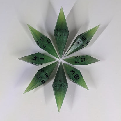 A complete set of resin dice featuring 2 d20s for advantage rolls. These swirling shades of green with black numbering slender handmade diamond dice are clustered in a tight circle on a white background, highlighting the quality of the artisan dice craftsmanship and the unique pointed shape.