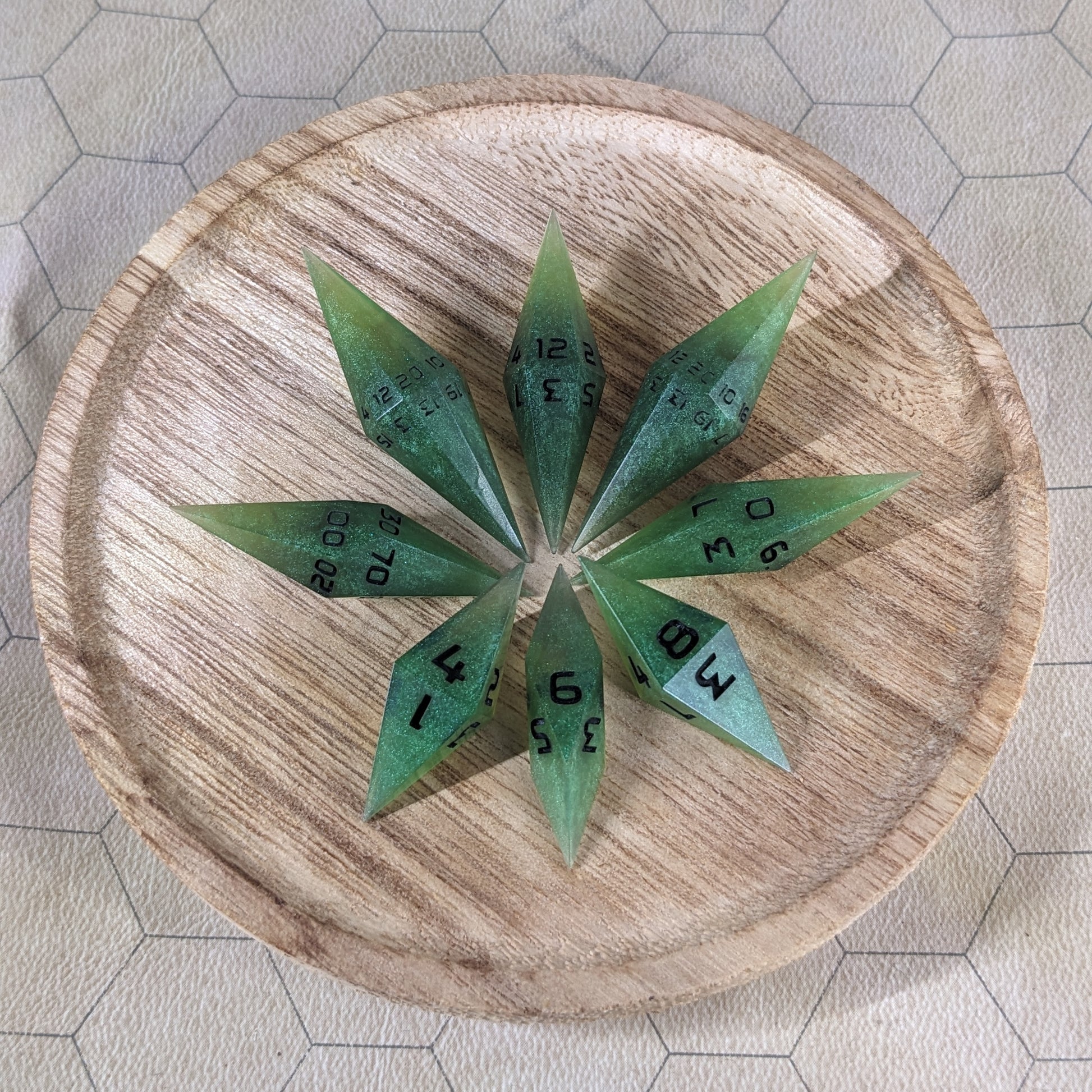 A set of swirling shades of green with black numbering handmade dice arranged in a wooden circle organizer. The resin dice feature a unique pointed diamond shape with the tips facing outward, highlighting the sharp edges of the two d20 and other artisan dice in the collection. 