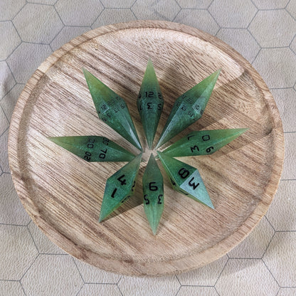 A set of swirling shades of green with black numbering handmade dice arranged in a wooden circle organizer. The resin dice feature a unique pointed diamond shape with the tips facing outward, highlighting the sharp edges of the two d20 and other artisan dice in the collection. 