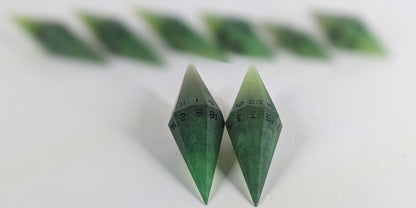 A stylized macro shot focusing heavily on 2 d20s from a swirling shades of green with black numbering handmade dice set in the foreground. The sharp diamond edges of these front resin dice are crisp, while the rest of the matching artisan dice collection forms a softly blurred background, creating depth.