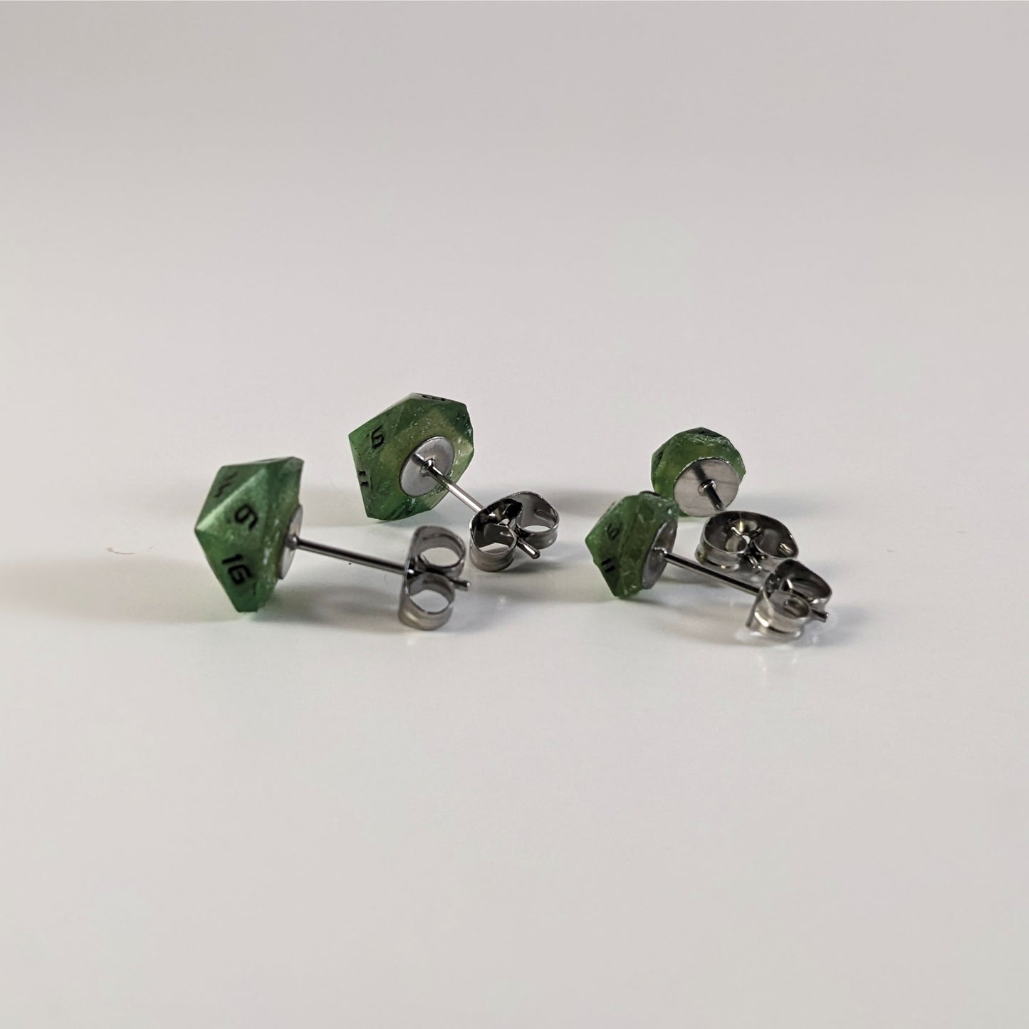 Two pairs of handmade d20 earrings that are shimmering green with black numbers turned to show the metal alloy hardware and butterfly backs.
