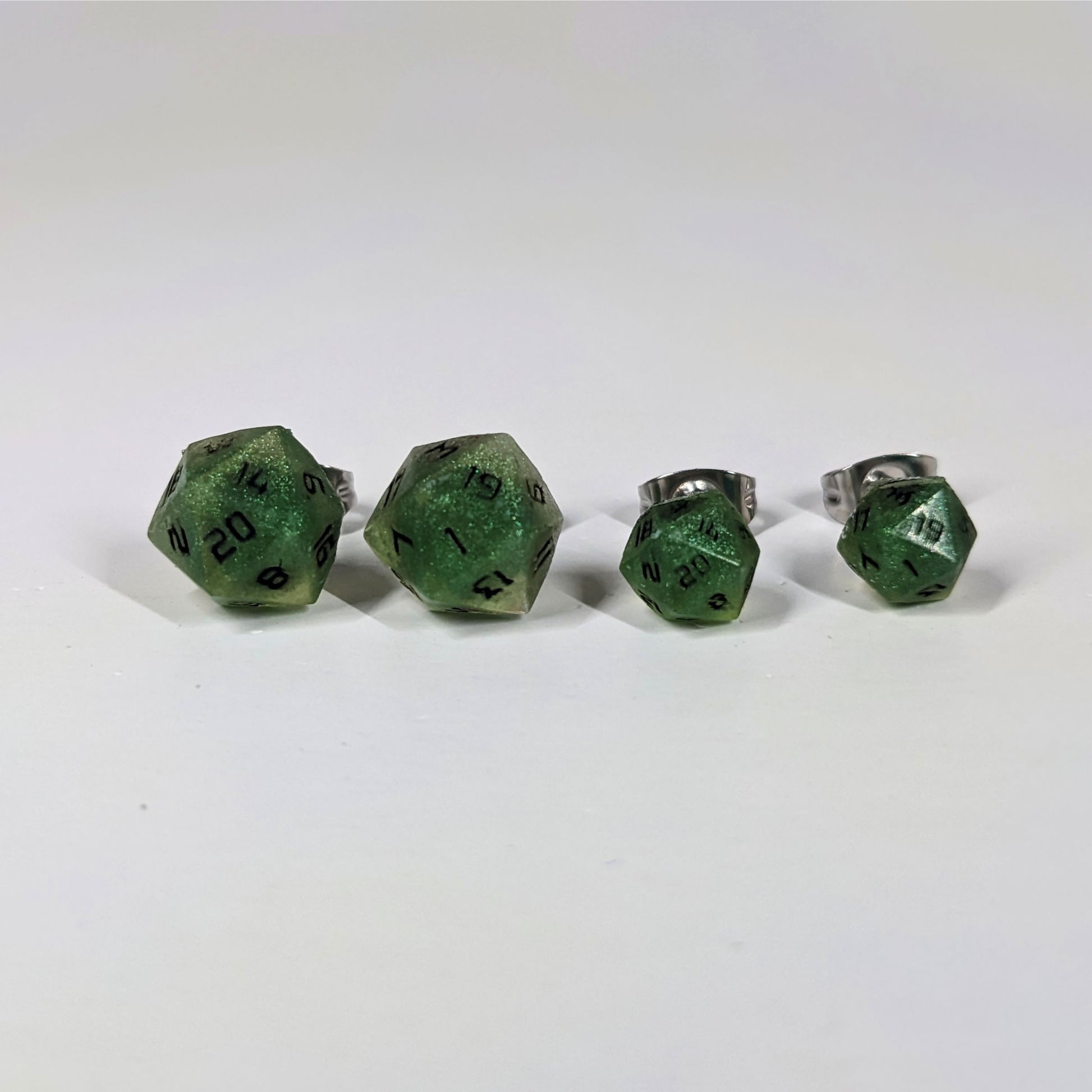 Two pairs of handmade d20 earrings that are shimmering green with black numbers on a white background sit in a line with the larger pair on the left and the smaller pair on the right. The earrings on the left of each pair centers the 20 face and the earrings on the right of each pair centers the 1 face.