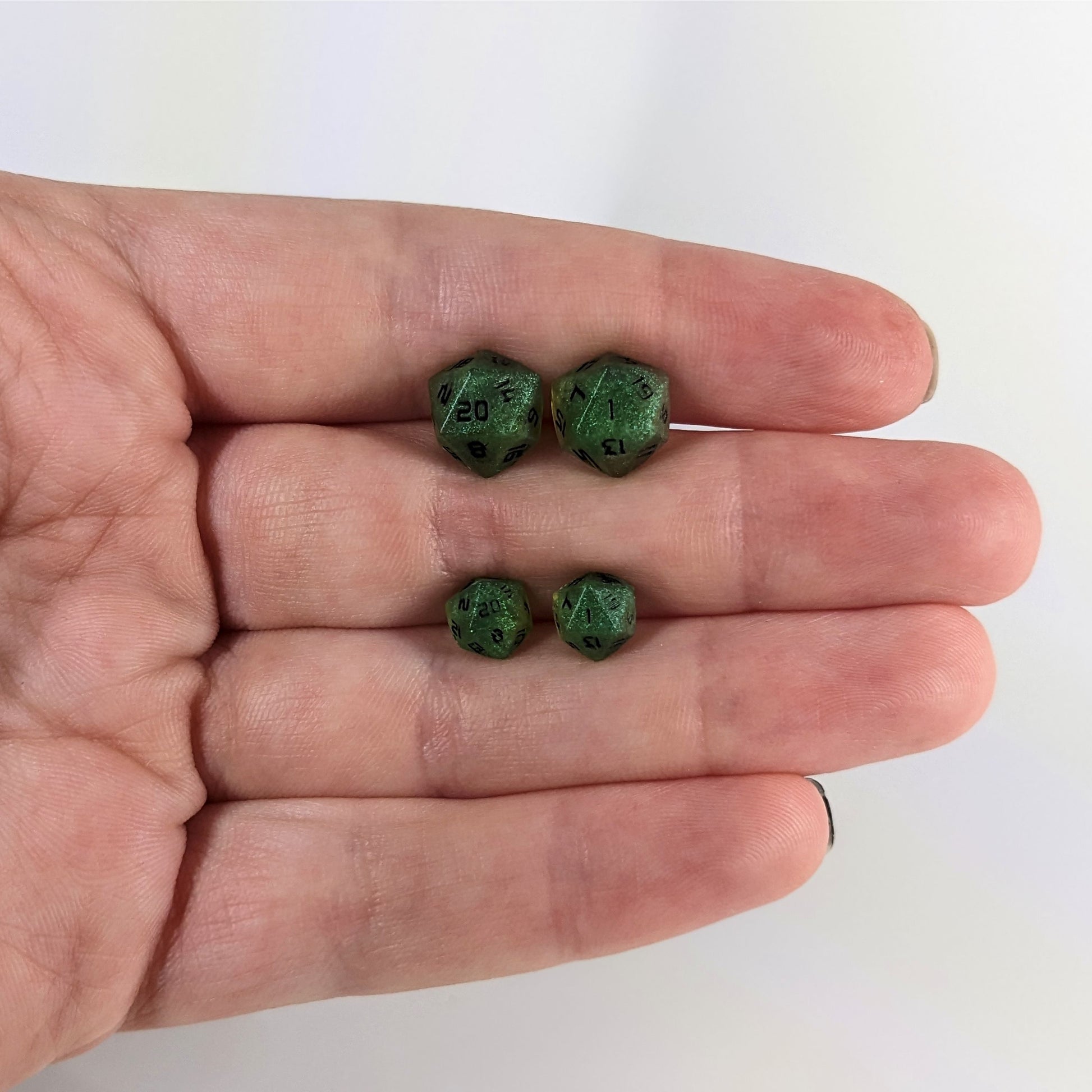 Two pairs of handmade d20 earrings that are shimmering green with black numbers held between the fingers of a flat hand. The larger pair is between the pointer and middle finger, the smaller between the middle and ring finger. The earrings on the left of each pair centers the 20 face and the earrings on the right of each pair centers the 1 face.