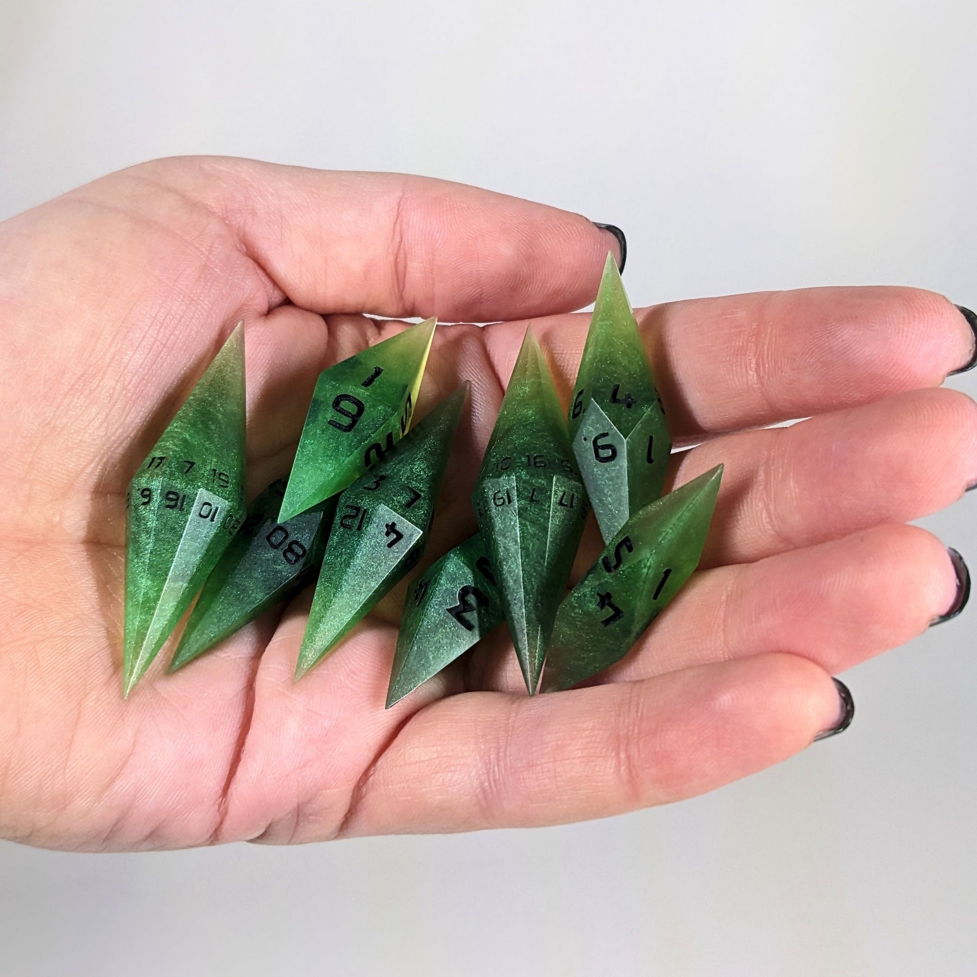 An 8 dice set of diamond style dice that are swirling shades of green with black numbers are held in a hand. The image shows that the set fits easily in a hand, making them overall smaller, though sharper, than a standard set of dnd dice.