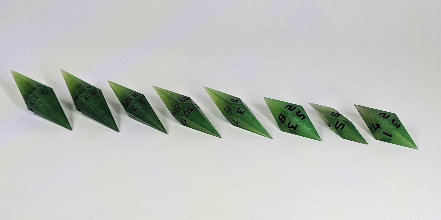 A mid angle photo of a complete set of handmade diamond DnD dice that are swirling shades of green with black numbers arranged in a straight line with bottom points angled slightly forwards and towards the right. They are lined up with the 2 d20s on the left going down to the d4 on the right side. The highest value number of each die is facing forward.