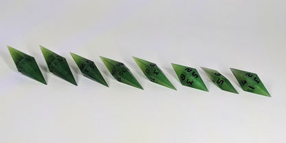A mid angle photo of a complete set of handmade diamond DnD dice that are swirling shades of green with black numbers arranged in a straight line with bottom points angled slightly forwards and towards the right. They are lined up with the 2 d20s on the left going down to the d4 on the right side. The highest value number of each die is facing forward.