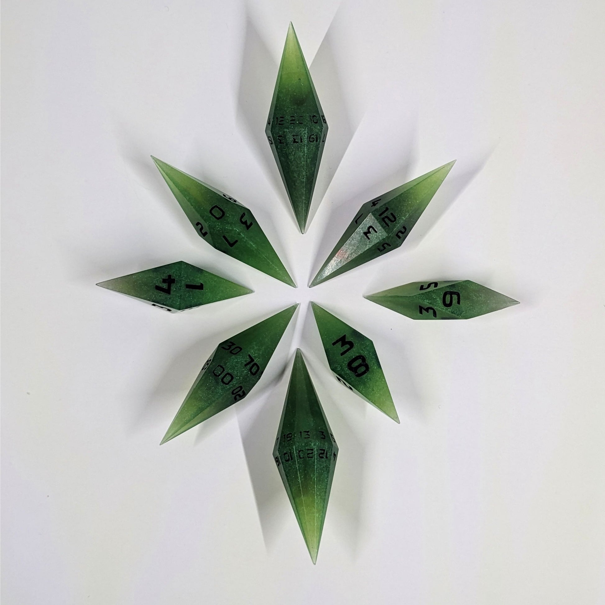 A complete set of diamond shaped resin dice featuring 2 d20s. These handmade dice are swirling shades of green with black numbers and are clustered in a circle with the bottom points angled toward a central point on a white background.