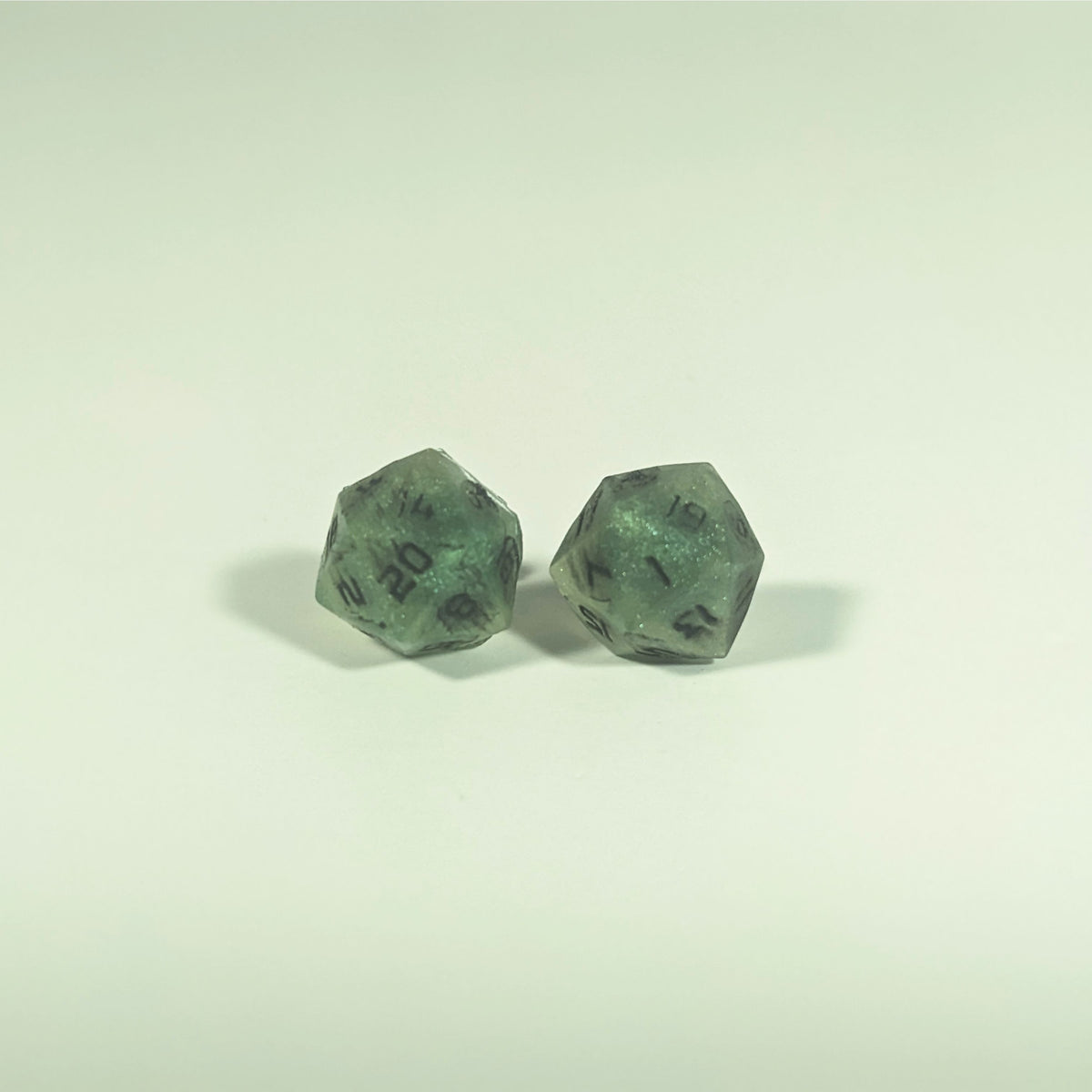 A single pair of the larger size handmade d20 earrings with titanium hardware that are swirls of green with black numbers on a white background. The earring on the left centers the 20 face and the earring on the right centers the 1 face.