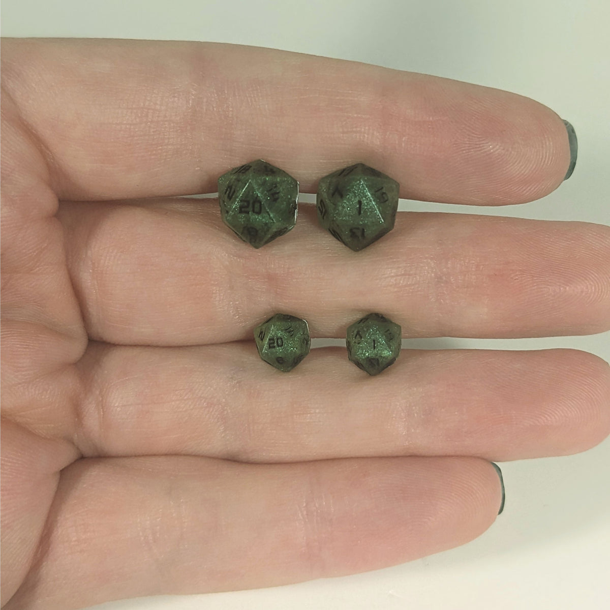 Two pairs of handmade d20 earrings with titanium hardware that are swirls of green with black numbers held between the fingers of a flat hand. The larger pair is between the pointer and middle finger, the smaller between the middle and ring finger. The earrings on the left of each pair centers the 20 face and the earrings on the right of each pair centers the 1 face.
