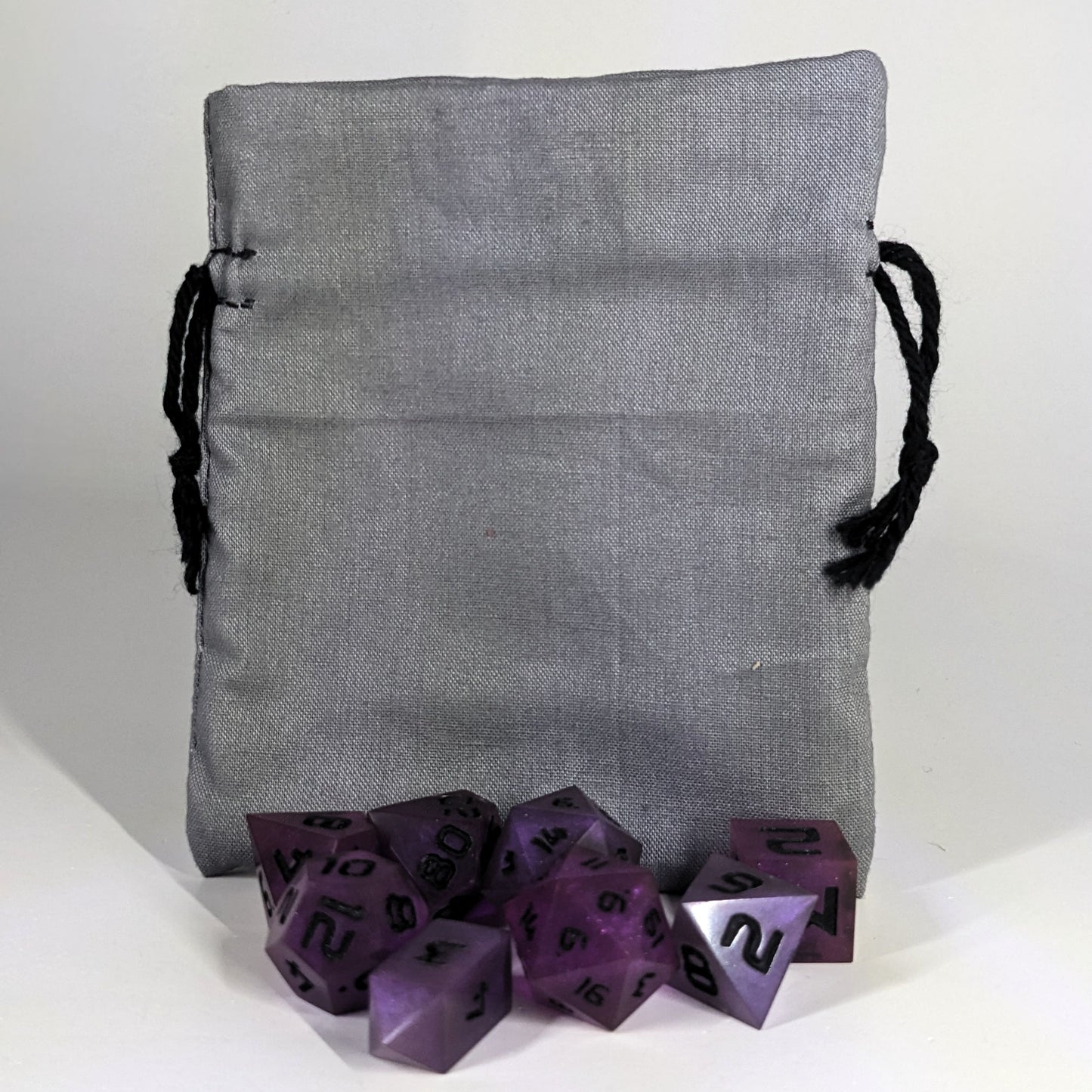 A small, handmade dice bag that is gray stands upright on a white background with a set of purple dnd dice with black numbers sitting in front of it.