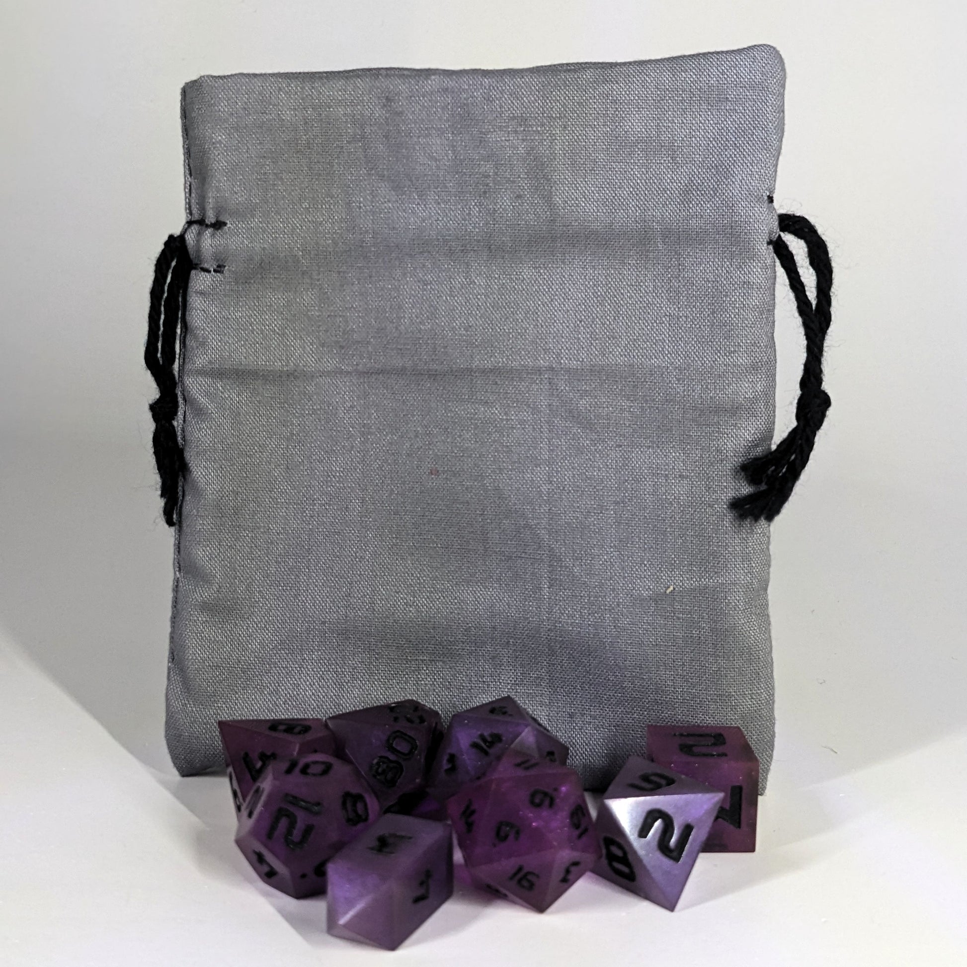A small, handmade dice bag that is gray stands upright on a white background with a set of purple dnd dice with black numbers sitting in front of it.
