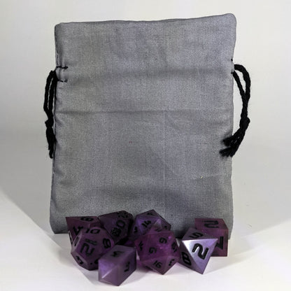 A small, handmade dice bag that is gray stands upright on a white background with a set of purple dnd dice with black numbers sitting in front of it.