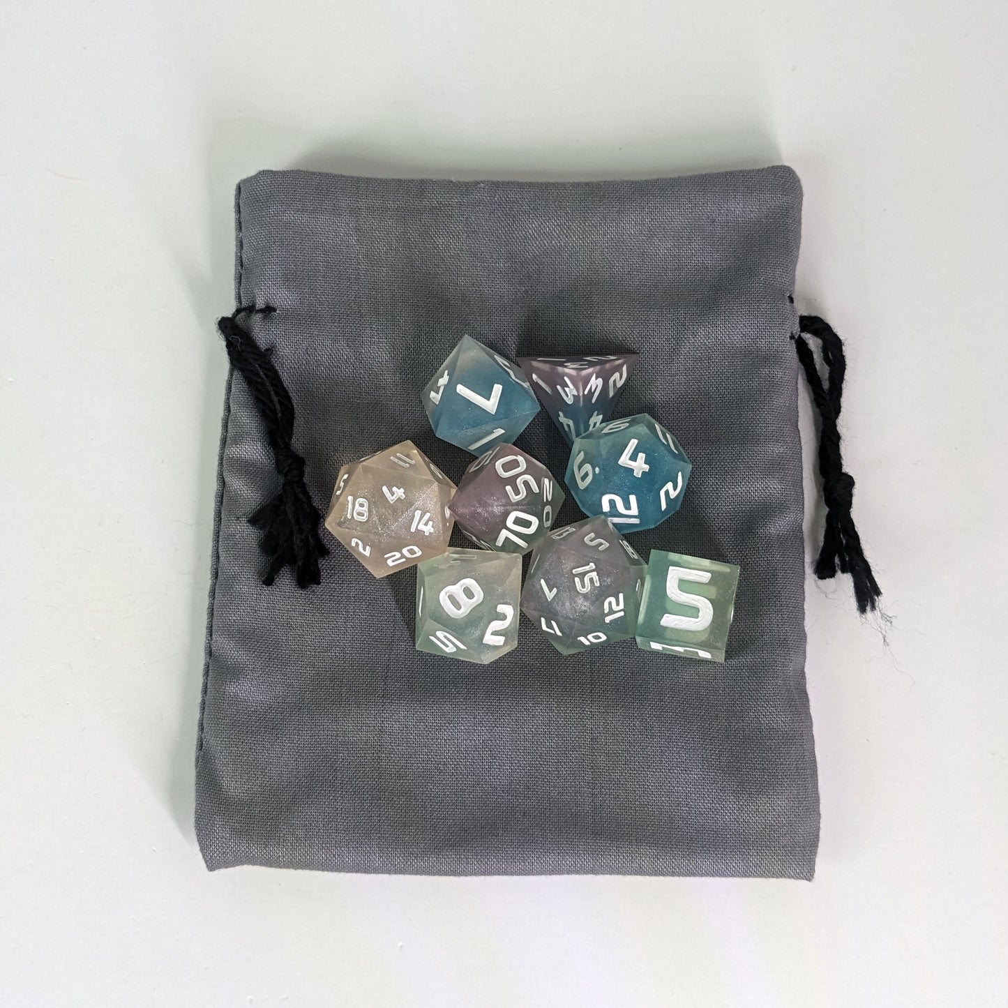 A small, handmade dice bag that is gray lays flat on a white background with a set of colorful dnd dice with white numbers sitting on top of it.