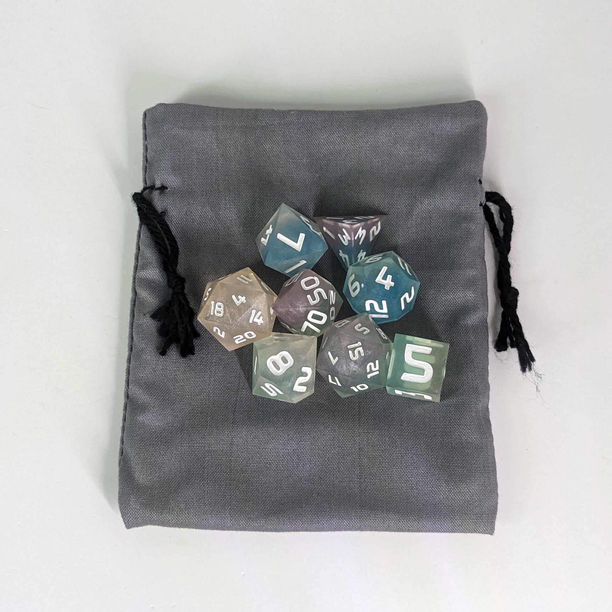 A small, handmade dice bag that is gray lays flat on a white background with a set of colorful dnd dice with white numbers sitting on top of it.