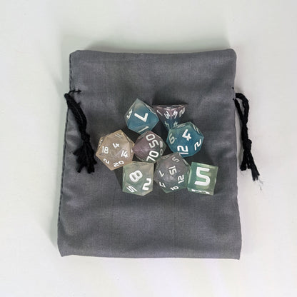 A small, handmade dice bag that is gray lays flat on a white background with a set of colorful dnd dice with white numbers sitting on top of it.