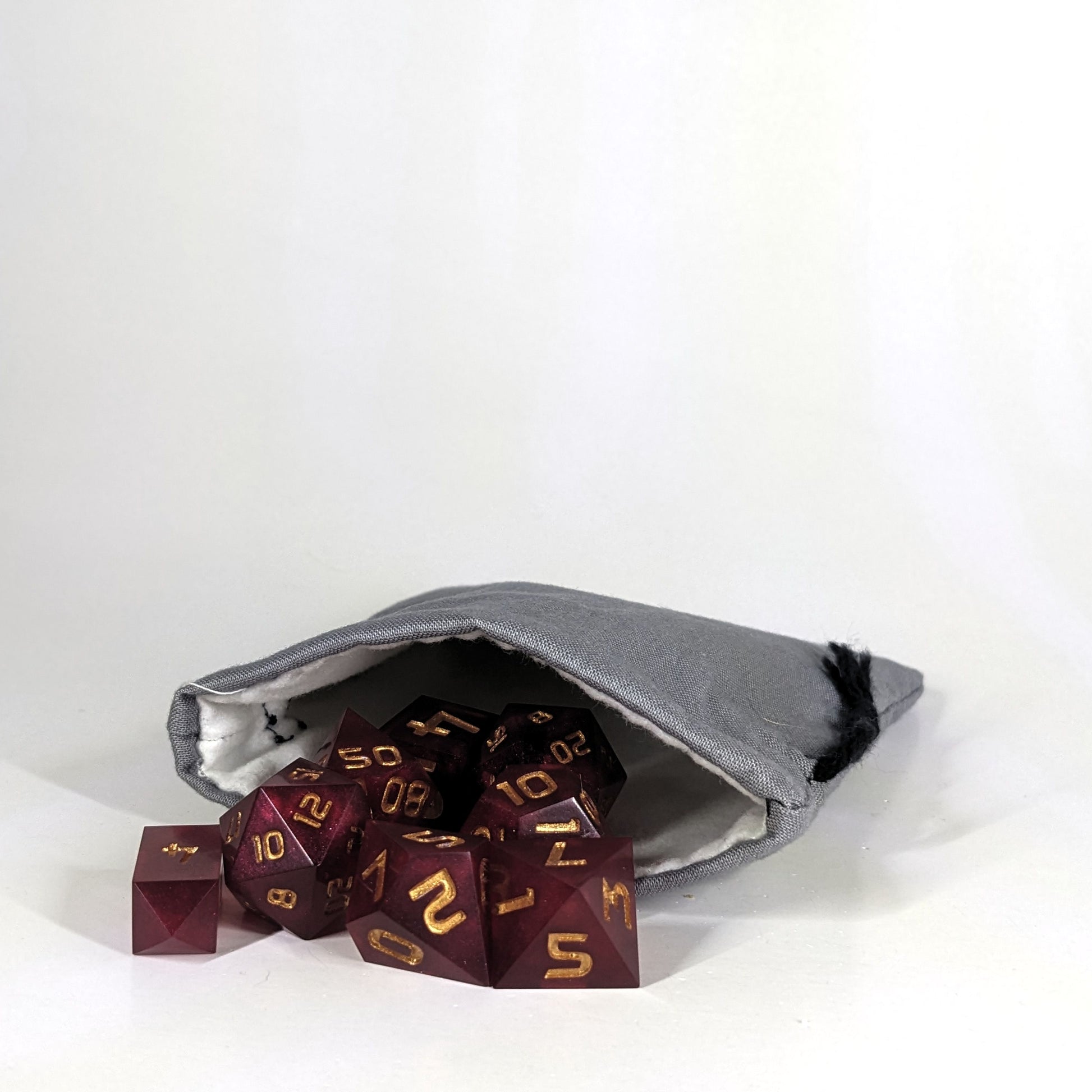 A small, handmade dice bag that is gray lays open with a set of red, dnd dice with gold numbers spilling out of it on a white background.
