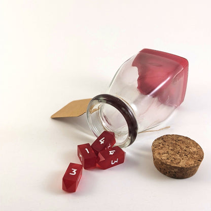 A healing potion bottle is tipped over with dice spilling out of it on a white background. The cork stopper is to the right of the bottle opening, and the blank, kraft tag is hanging off the left side of the bottle. The layer of red resin at the bottom of the bottle shows that it is certainly not liquid, and four small red crystal d4s appear to spill out the bottle opening.