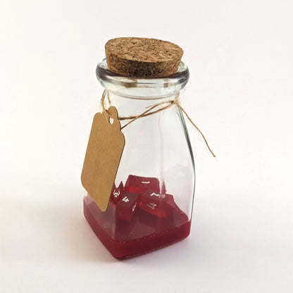 A healing potion bottle sits closed on a white background. The cork stopper is closing the top, a blank kraft color tag is hanging around the neck. Inside the bottle are 4 small red crystal d4s and a layer of red resin.
