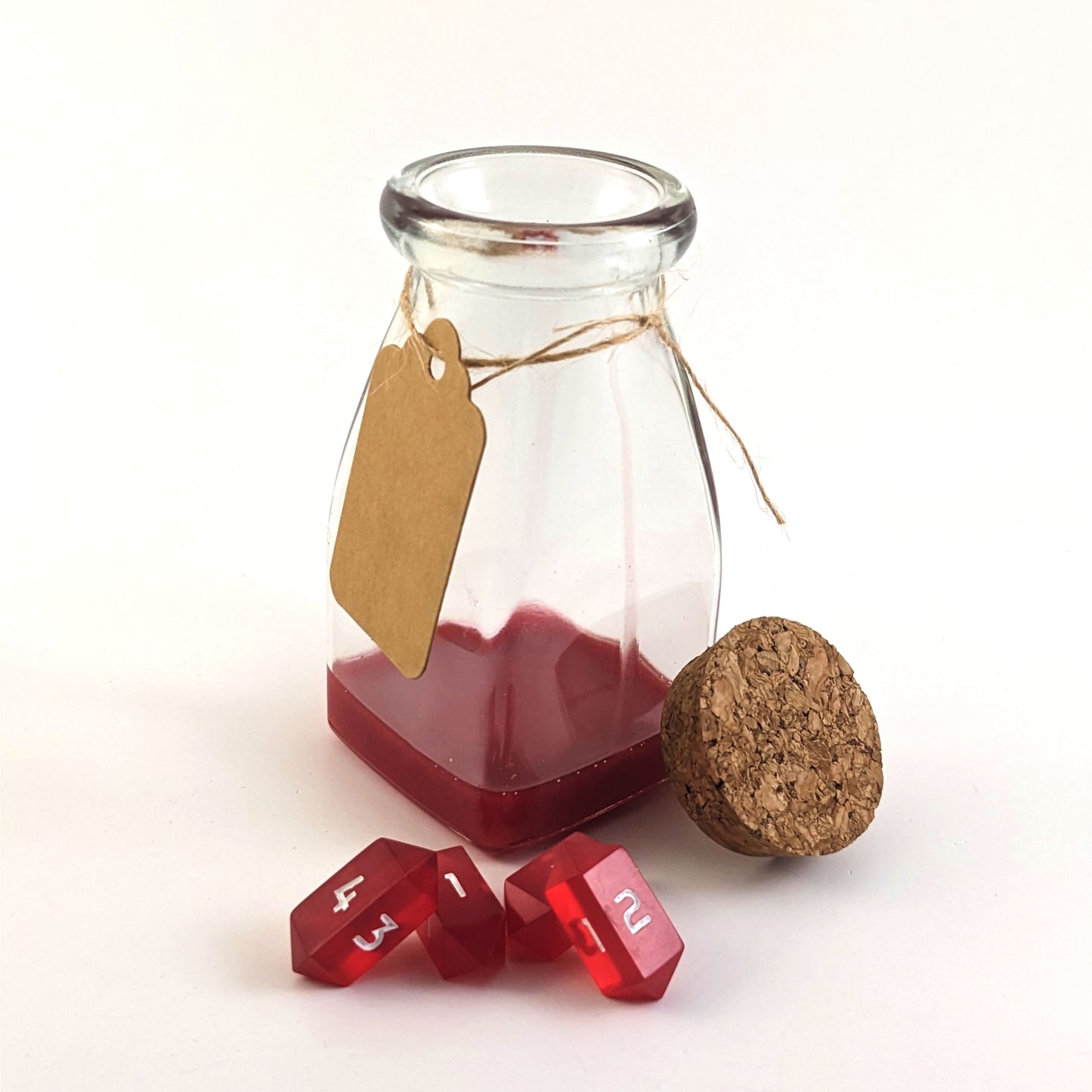 A healing potion bottle sits open with dice in front of it on a white background. The cork stopper is propped up on the glass bottle on the left side, and on the right the blank, kraft tag hangs around the bottle neck. There is a layer of red resin on the bottom of the bottle, and four small red crystal d4s are piled in front of the bottle.