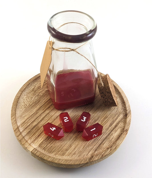 A glass bottle sits open on a wooden circle. The cork stopper rests propped up on the side of the glass bottle. Inside the bottle is red resin to look like liquid healing potion. Four small red crystal d4s rest in front of the bottle in an arc with the 4, 2, 3, and 1 faces up from left to right.