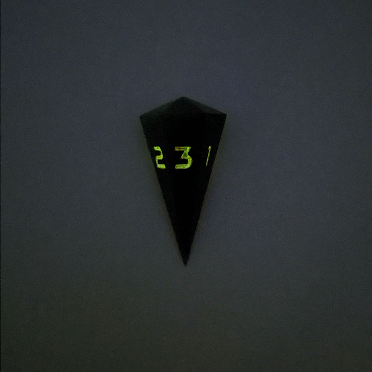 A high angle view of a dagger d3 with swirls of black and green with orange numbers that glow in the dark on a white background. This photo is taken in the dark to show the black numbers glowing green in the dark.