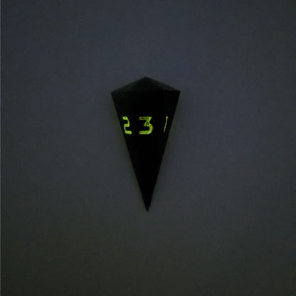 A high angle view of a dagger d3 with swirls of black and green with orange numbers that glow in the dark on a white background. This photo is taken in the dark to show the black numbers glowing green in the dark.
