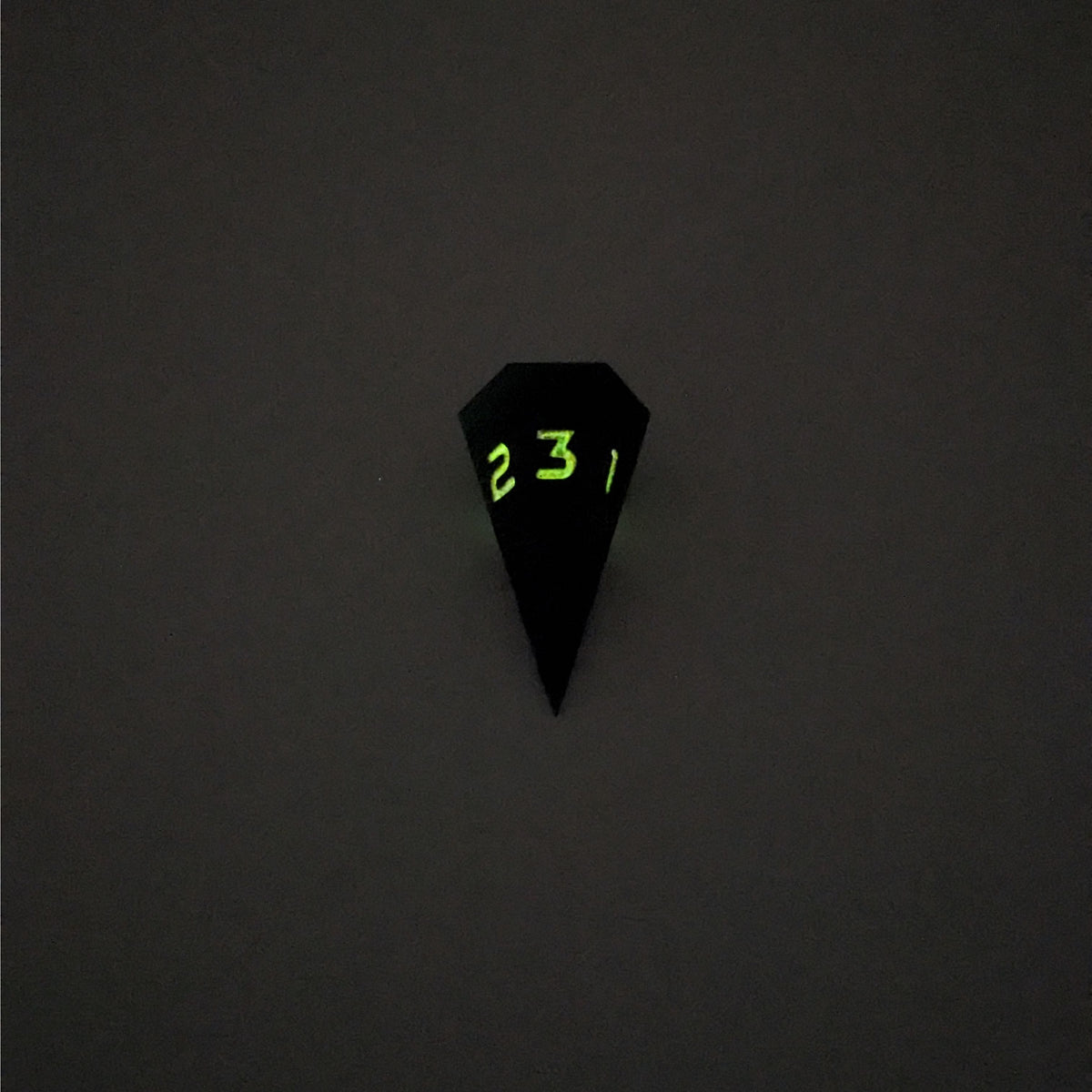 A mid angle view of a dagger d3 with swirls of black and green with orange numbers that glow in the dark on a white background. This photo was taken in the dark to show the black numbers glowing green in the dark.