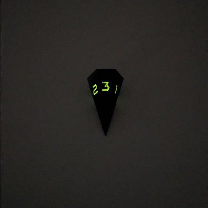 A mid angle view of a dagger d3 with swirls of black and green with orange numbers that glow in the dark on a white background. This photo was taken in the dark to show the black numbers glowing green in the dark.