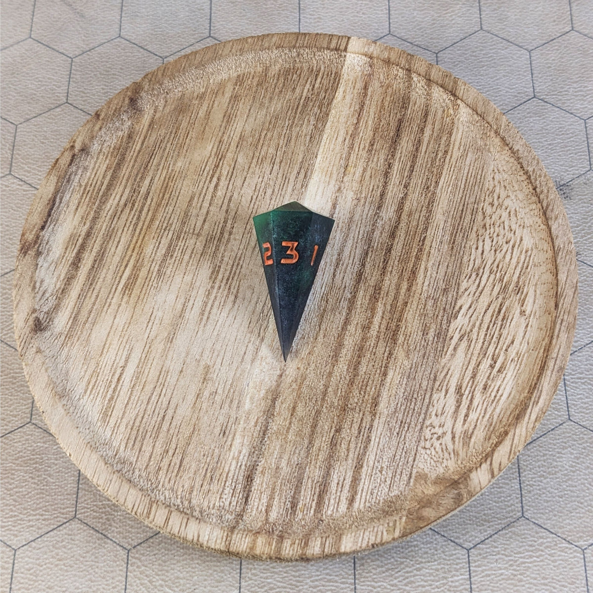 A high angle view of a dagger d3 with swirls of black and green with orange numbers that glow in the dark. It is sitting in the center of a wooden circle.