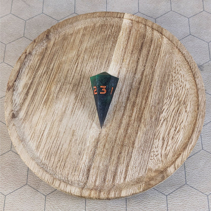 A high angle view of a dagger d3 with swirls of black and green with orange numbers that glow in the dark. It is sitting in the center of a wooden circle.