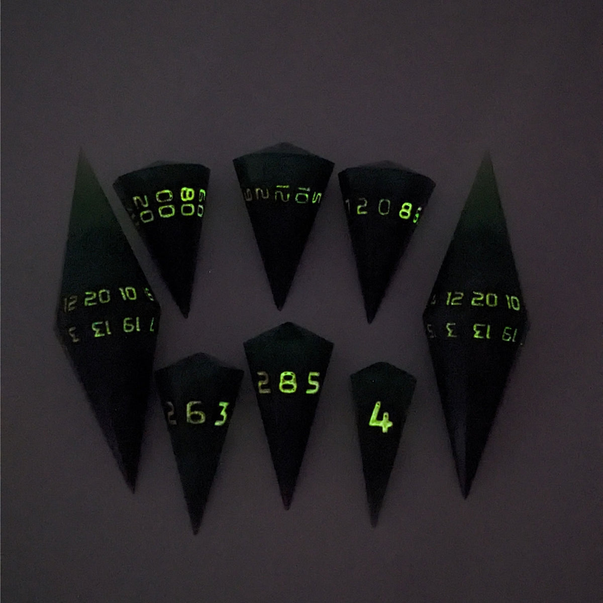 A dagger style DnD dice set that is  green and black with orange numbers that glow in the dark is arranged with a d20 each on the right and left, with the d%, d12, and d10 along the top and the d6, d8, and d4 along the bottom, all in line with the d20s. The photo was taken in the dark to show the orange numbers glowing green.