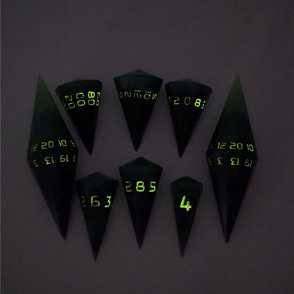 A dagger style DnD dice set that is  green and black with orange numbers that glow in the dark is arranged with a d20 each on the right and left, with the d%, d12, and d10 along the top and the d6, d8, and d4 along the bottom, all in line with the d20s. The photo was taken in the dark to show the orange numbers glowing green.