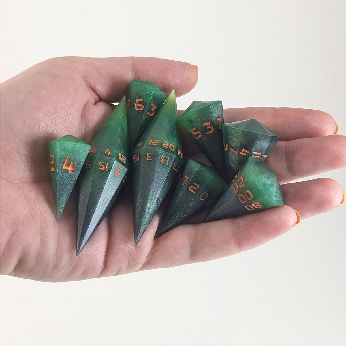 An 8 dice set of dagger style dice that are green and black with orange numbers that glow in the dark are held in a hand. The image shows that the set just fits in a hand, making them overall larger than a standard set of dnd dice.