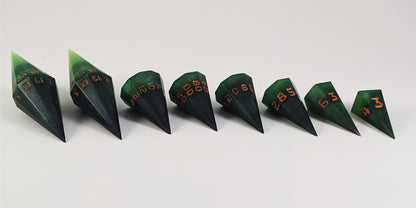 A straight on photo of a complete set of dagger DnD dice that are black and green with orange numbers that glow in the dark arranged in a straight line with points angled toward the right. They are lined up with the 2 d20s on the left going down to the d4 on the right side. The highest value number of each die is facing forward.