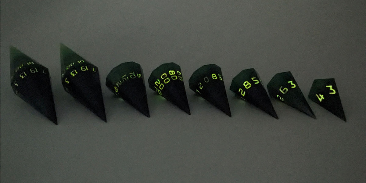 A straight on photo of a complete set of dagger DnD dice that are black and green with orange numbers that glow in the dark arranged in a straight line with points angled toward the right. They are lined up with the 2 d20s on the left going down to the d4 on the right side. The highest value number of each die is facing forward. The photo is taken in the dark to show the orange numbers glowing green.