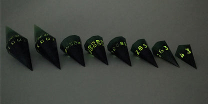 A straight on photo of a complete set of dagger DnD dice that are black and green with orange numbers that glow in the dark arranged in a straight line with points angled toward the right. They are lined up with the 2 d20s on the left going down to the d4 on the right side. The highest value number of each die is facing forward. The photo is taken in the dark to show the orange numbers glowing green.