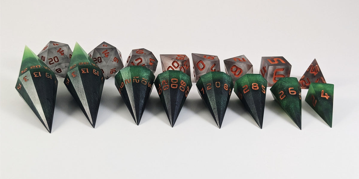 A straight on photo of a complete set of dagger dice that are green and black with orange numbers that glow in the dark arranged in a straight line with points angled forward. They are lined up with the 2 d20s on the left going down to the d4 on the right side. Behind the dagger set is a standard set of polyhedral dnd dice to allow for size comparison. The dagger dice are much larger overall, but not taller. The highest value number of each dagger die is facing up.