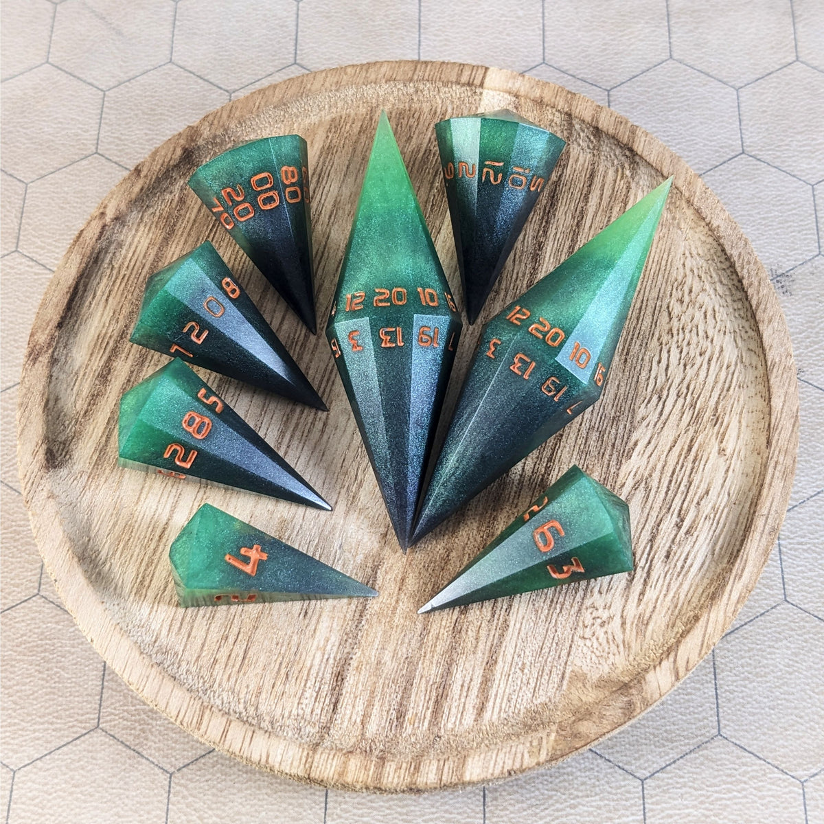 A complete set of dagger style resin dice that are green and black with orange numbers that glow in the dark are arranged on a wooden circle. The 2 d20s are next to each other with the d12 placed between them on top. The d6 is to the bottom right of the d20s and the rest of the dice are arranged along the left side, points angled toward the d20s.