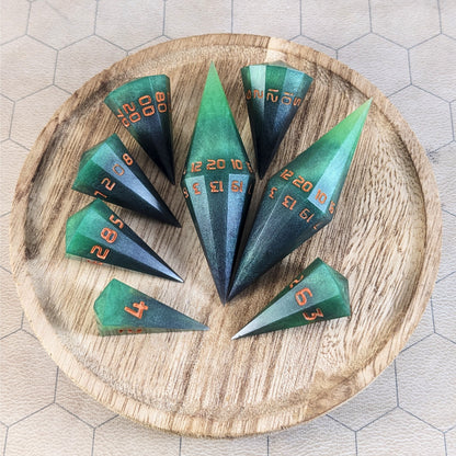 A complete set of dagger style resin dice that are green and black with orange numbers that glow in the dark are arranged on a wooden circle. The 2 d20s are next to each other with the d12 placed between them on top. The d6 is to the bottom right of the d20s and the rest of the dice are arranged along the left side, points angled toward the d20s.