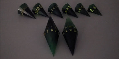 A straight on photo of a dagger set of DnD dice that are green and black with orange numbers that glow in the dark focusing on 2 d20s in the center. The d20 on the left has the 1 face up and the d20 on the right has the 20 face up. The other 6 dice are lined up in the background but are out of focus, while the d20s are in sharp focus. The photo is taken in the dark to show the orange numbers glowing green.
