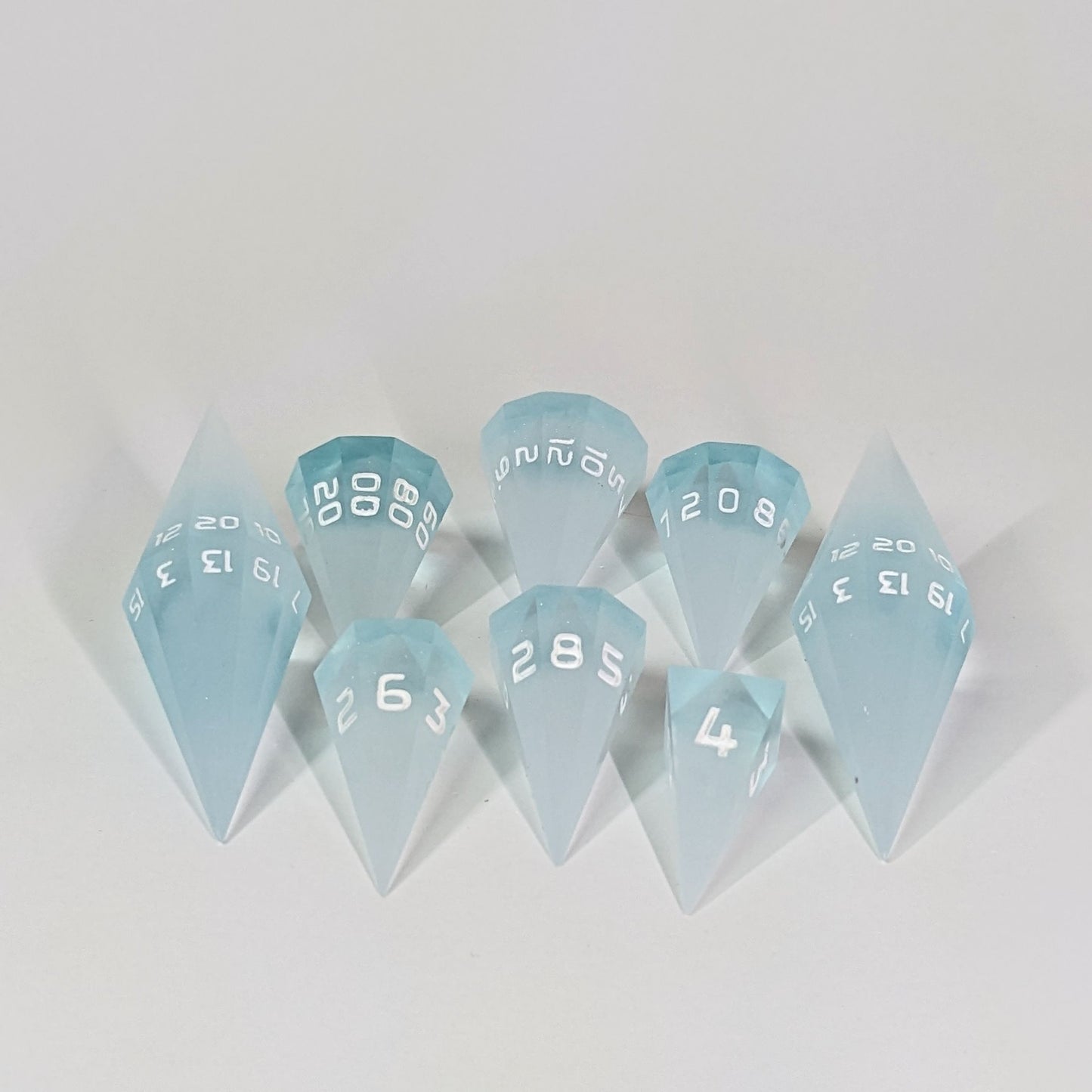 A dagger style DnD dice set that is frosty blue and white with a touch of sparkle with opaque white numbers is arranged with a d20 each on the right and left, with the d%, d12, and d10 along the top and the d6, d8, and d4 along the bottom, all in line with the d20s.