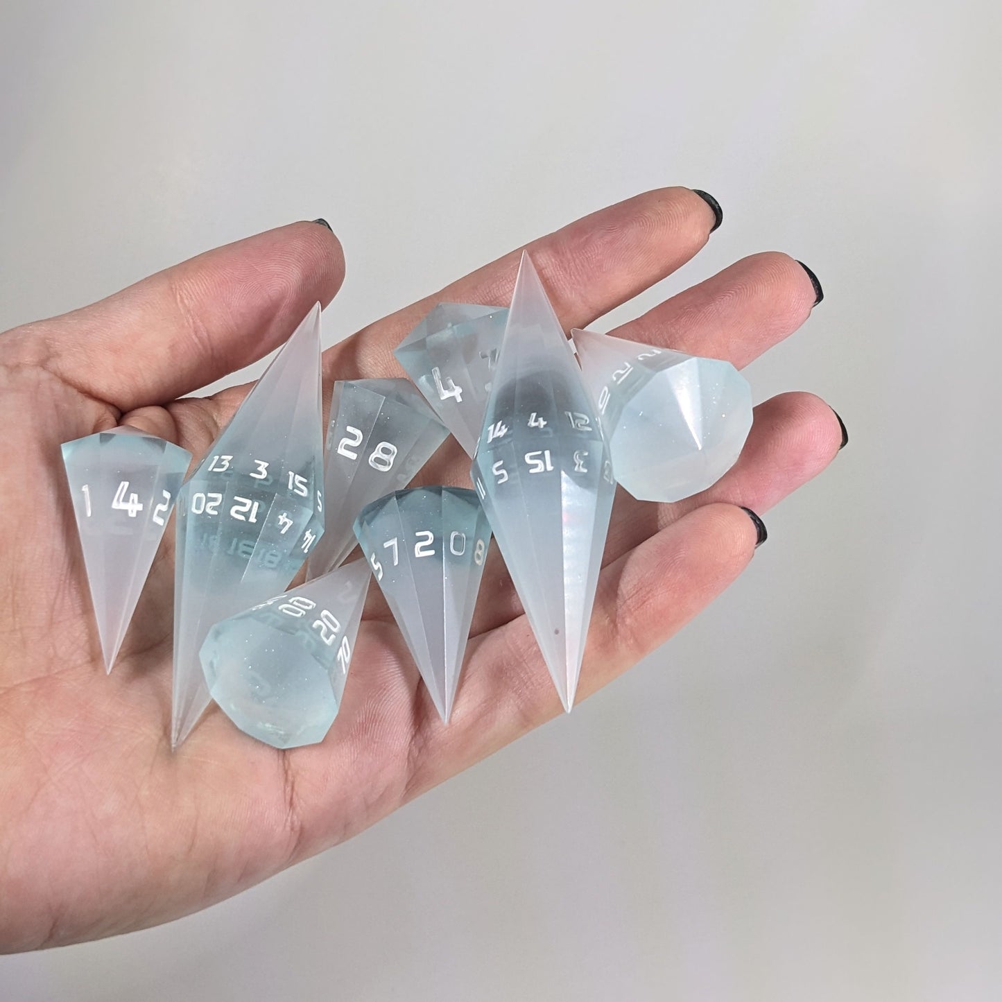 An 8 dice set of dagger style dice that are frosty blue and white with a touch of sparkle with opaque white numbers are held in a hand. The image shows that the set just fits in a hand, making them overall larger than a standard set of dnd dice.