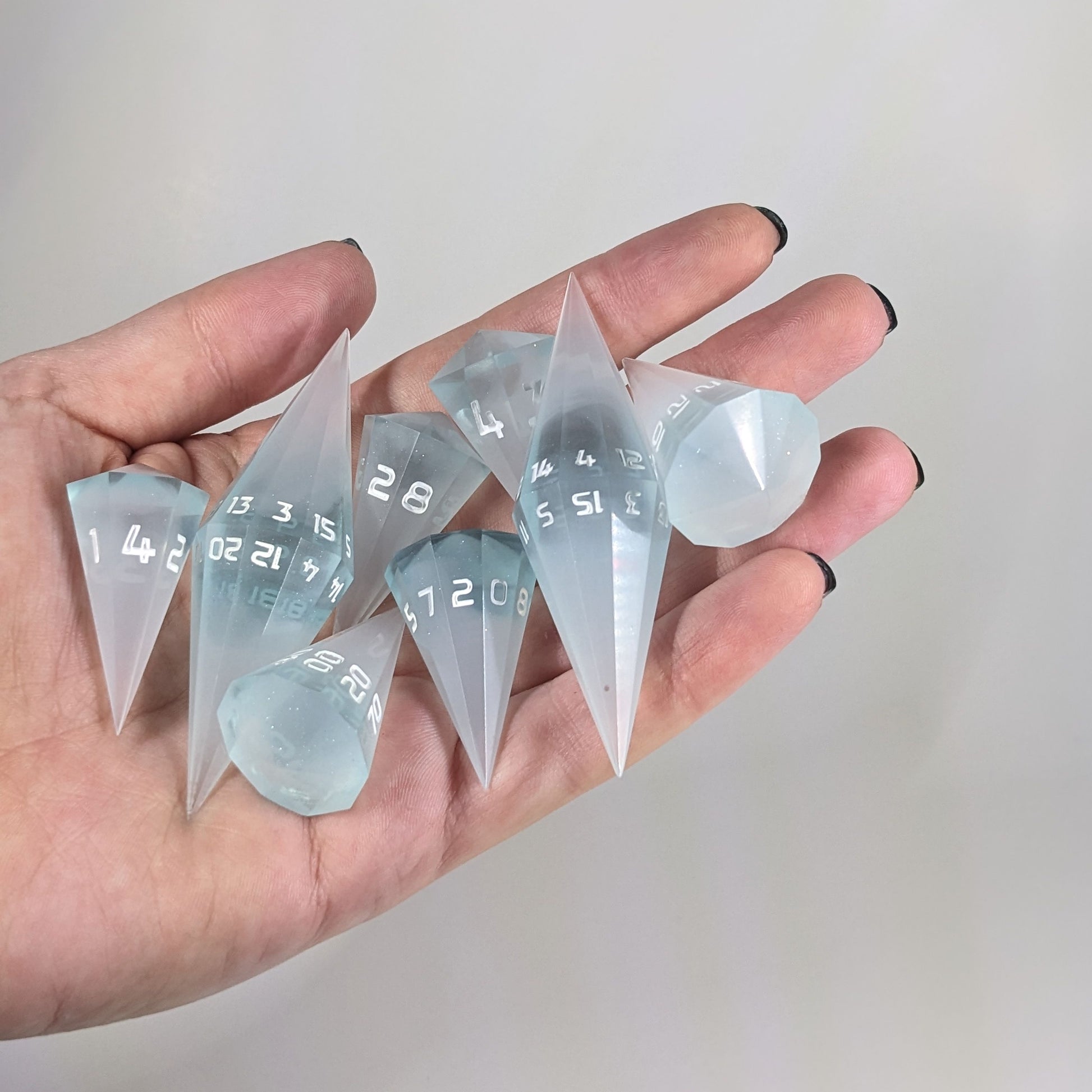 An 8 dice set of dagger style dice that are frosty blue and white with a touch of sparkle with opaque white numbers are held in a hand. The image shows that the set just fits in a hand, making them overall larger than a standard set of dnd dice.
