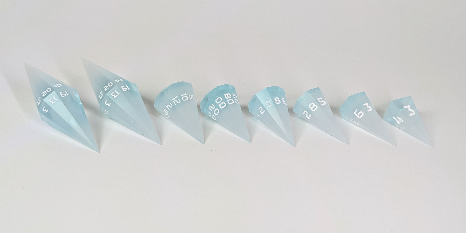A straight on photo of a complete set of dagger DnD dice that are frosty blue and white with a touch of sparkle with opaque white numbers arranged in a straight line with points angled toward the right. They are lined up with the 2 d20s on the left going down to the d4 on the right side. The highest value number of each die is facing forward.