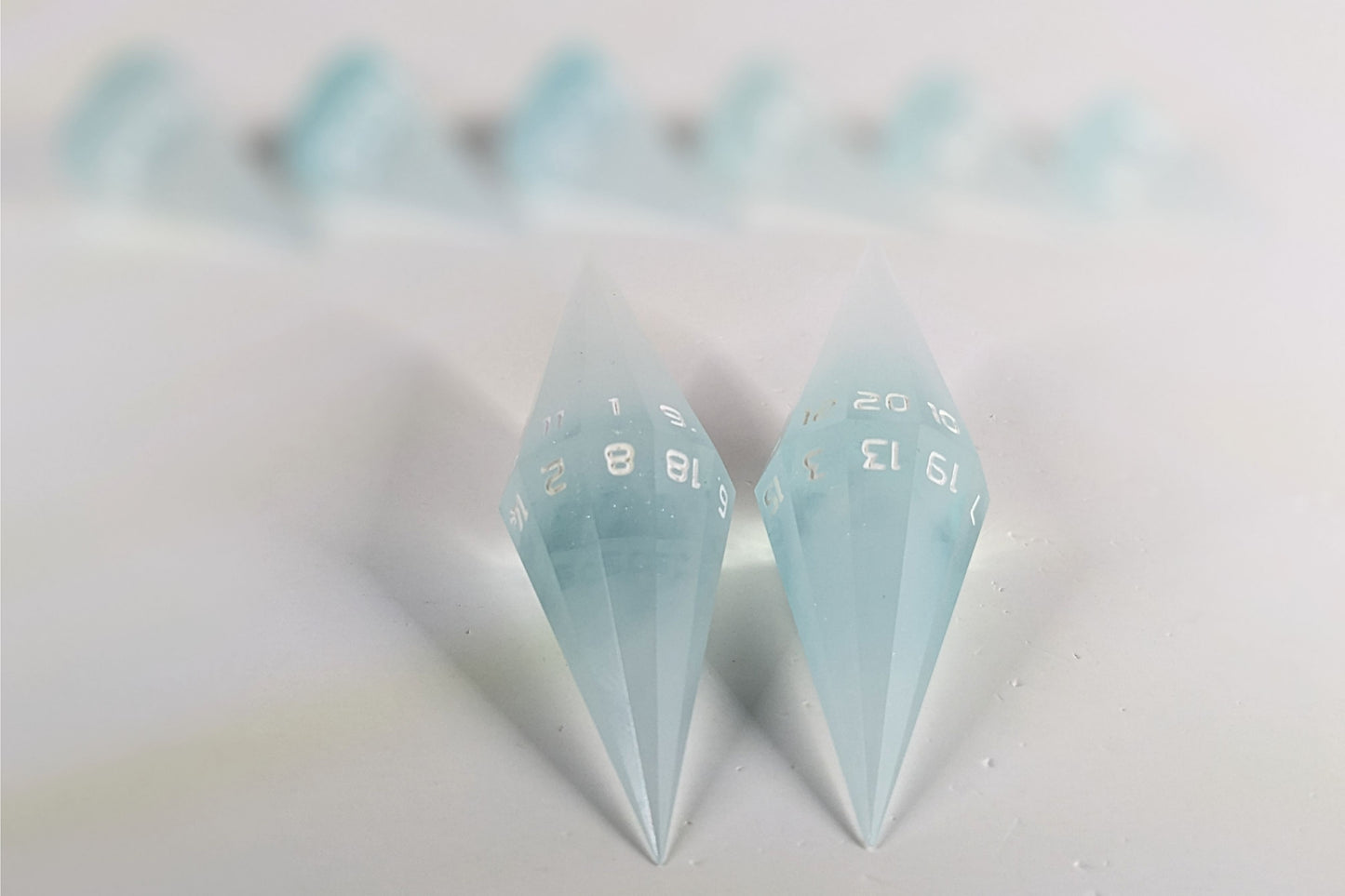 A straight on photo of a dagger set of DnD dice that are frosty blue and white with a touch of sparkle with opaque white numbers focusing on 2 d20s in the center. The d20 on the left has the 1 face up and the d20 on the right has the 20 face up. The other 6 dice are lined up in the background but are out of focus, while the d20s are in sharp focus.
