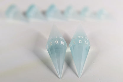 A straight on photo of a dagger set of DnD dice that are frosty blue and white with a touch of sparkle with opaque white numbers focusing on 2 d20s in the center. The d20 on the left has the 1 face up and the d20 on the right has the 20 face up. The other 6 dice are lined up in the background but are out of focus, while the d20s are in sharp focus.