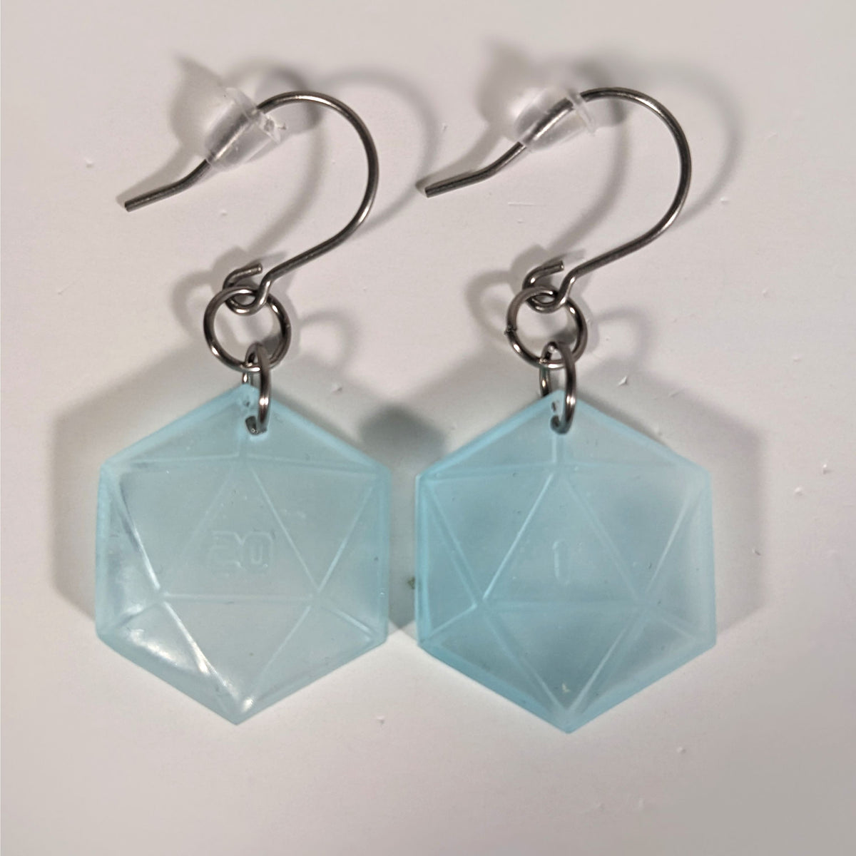 A pair of frosty blue and white dangly, flat d20 earrings with silver color titanium and niobium hardware and bell shaped rubber backs on a white background. The earring on the left has a 20 on it and the earring on the right has a 1 on it, both in a sci-fi style font.