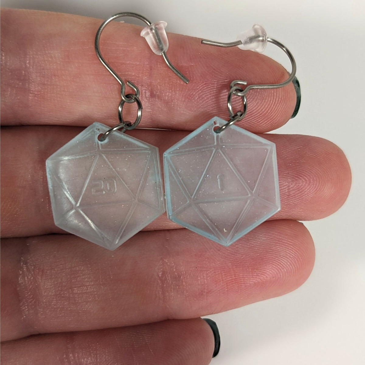 A pair of frosty blue and white dangly, flat d20 earrings with silver color titanium and niobium hardware with bell shaped rubber backs held on on a hand on a white background. The earring on the left has a 20 on it and the earring on the right has a 1 on it, both in a sci-fi style font.